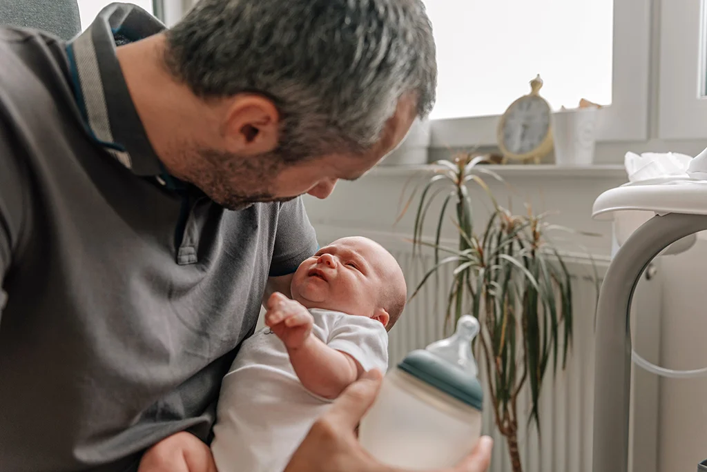 Father holding a newborn baby at home while preparing a bottle for feeding.