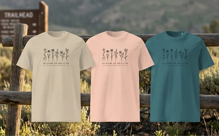 Three Trailhead Chiropractic merch T-shirts featuring the Bloom in Health design in neutral, pink, and teal colors.